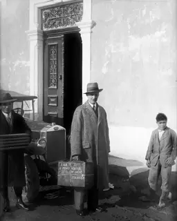 Three men and a boy outside a doorway; the central man in a tweed overcoat holds a lettered box beside a car.