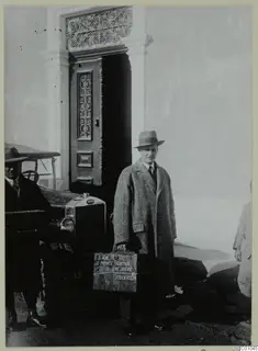 Einar Gjerstad in a tweed overcoat and hat, carrying a box, beside a car and a carved wooden doorway in Nicosia.