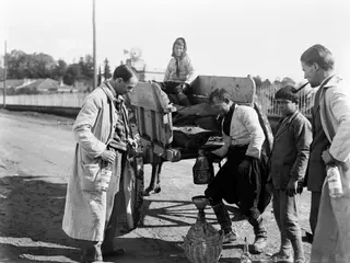 A man pours from a metal jug into a wicker-covered demijohn beside a cart, watched by a boy and two men in long coats.