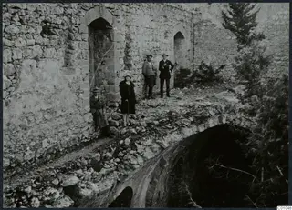Four people standing and sitting on a rubble ledge beside a ruined stone wall with arched openings, conifer trees at right