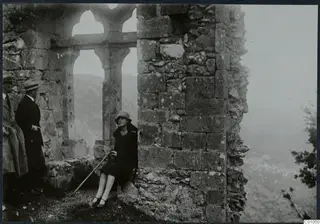 Two men stand beside a ruined stone arched window while a woman sits against a wall, with hills behind