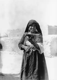 Woman in a dark headscarf and long dress holding a large painted vessel in front of a stone wall
