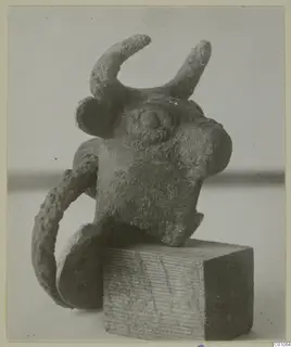 Bronze bull-head object with curved horns and a loop at one side, set on a wooden block
