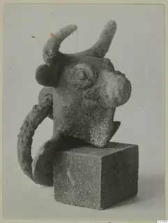 Bronze bull's head object with upturned horns and a curved loop, set on a square stone block
