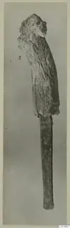 Bronze tool with a thick, crusted upper end and a long narrow flat blade, photographed upright against a grey background.