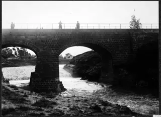 Brick bridge with two arches over fast-flowing water at Nicosia, with three people standing on its railed deck.