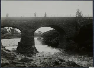 Brick bridge with two arches over fast-flowing water, with three people standing on its railing.