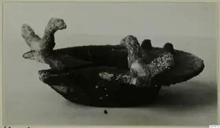 Corroded metal lamp with two upright projections, photographed from the side on a plain surface