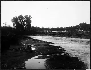 River with fast-flowing water, stony banks and a line of trees on the far bank, with a windmill at the left edge.