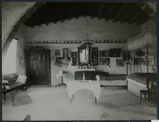 Interior of a whitewashed room with a canopied iron bed, a wardrobe, a round table and framed pictures on the wall, at Idalion.
