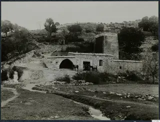 Stone building with a tall arched opening and a square tower at the Old Mill, Kalavasos, with two animals and several people