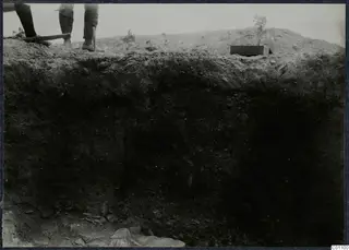 Vertical section of dark excavated soil at Idalion Acropolis, with the legs of two people and a tool handle at the top edge.