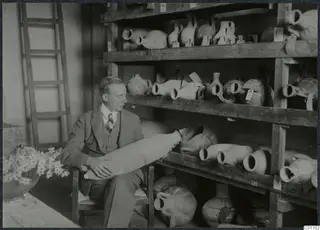 John Lindros seated with a large pottery vessel on his lap, beside shelves of jugs and jars
