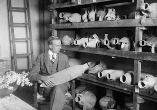 John Lindros seated in a chair holding a large pottery vessel, with shelves of jugs and jars behind him