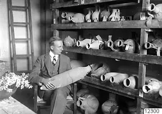 John Lindros seated with a large pottery vessel on his lap, in a room with shelves of pottery jars and jugs.
