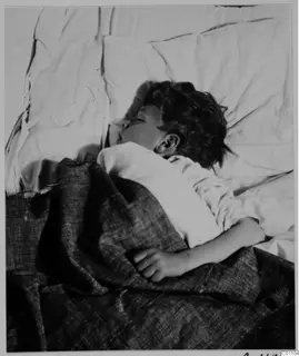 Sleeping child lying on a bed under a dark blanket, head resting on a white pillow.