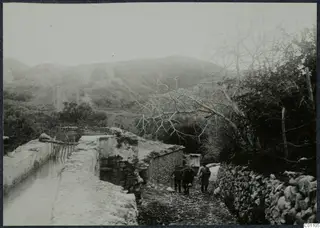 Water channel at Kythrea with two men walking up a stony path behind a laden donkey or mule.