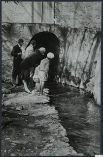 Visitors looking into a stream that flows from an arched opening in a stone wall at Kythrea