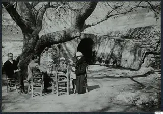 Group of people resting on wooden chairs under a bare tree beside a stone spring at Kythrea