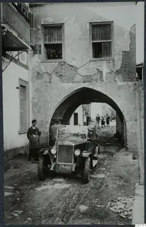 Open car driving along an unpaved street under an arch, with people watching from a barred window and standing nearby.
