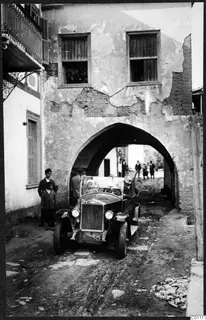 Open-topped car driving under a stone arch on a street in Kythrea, with people standing nearby.