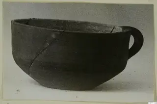 Pottery bowl with a single loop handle from the Kastros tombs at Lapithos, shown against a grey backdrop