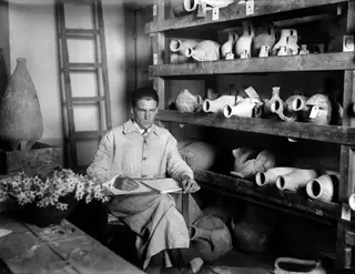 Alfred Westholm writing in a notebook beside wooden shelves of pottery jugs at the Swedish Institute, Nicosia.