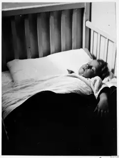 A child asleep on a pillow in a slatted wooden cot, one arm hanging over the edge