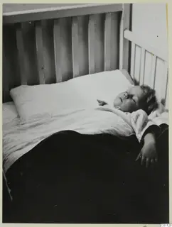 A young child asleep in a white-railed bed under a white pillow and dark blanket.