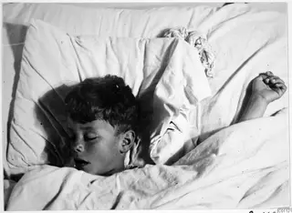 A young boy with dark hair asleep on a white pillow, one arm raised above the bedding.