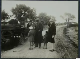 Two men, two children and a woman standing beside an open car on a dirt road at Idalion.