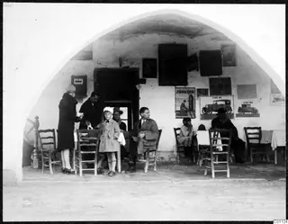 Several people seated and standing under a whitewashed arched veranda at Skarinou, with wooden chairs and tables with white cloths.
