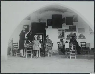 Seated and standing people outside a whitewashed arched café at Skarinou, with posters and café chairs
