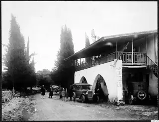 Cars and people standing in a dirt road outside a white two-storey building with arches and a wooden balcony, Skarinou