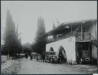 Group of people and two cars beside a two-storey white building with arches, on a road at Skarinou