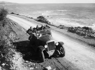 Open touring car with five people on a dusty road running along the coast beside the sea.
