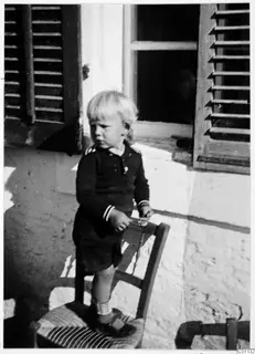 Young blond child standing on a chair against a whitewashed wall beside a shuttered window.