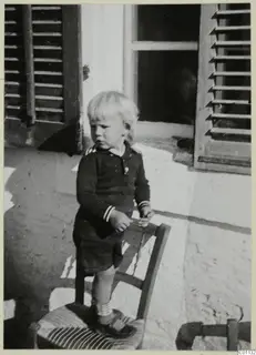 Small blond child in a dark sailor-style top standing on a wooden chair in front of a shuttered window.