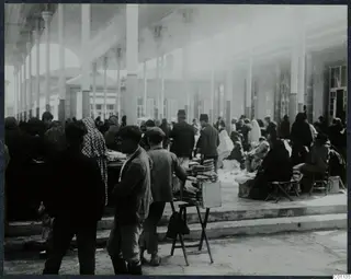 Covered market square at Nicosia, with tall columns, a crowd of people and a bread stall on a wooden stand.