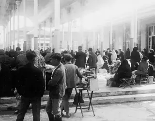 Market square in Nicosia with people in the shade of a colonnade, a vendor's stool stacked with bread rings in front.