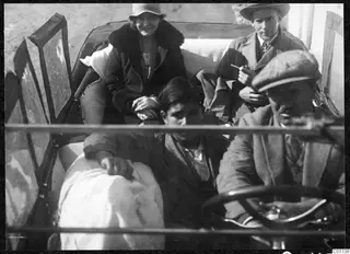 Two passengers in the back of an open car, and a man at the steering wheel with a boy sitting beside him.