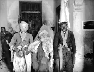 Three people in carnival costume stand in a whitewashed room, one with a cloth-wrapped head and one in shaggy fleece.
