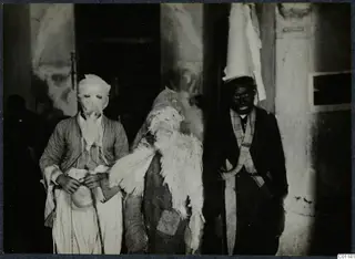 Three people in animal-skin and mask costumes stand together outside a doorway, last day of the carnival at Marion.