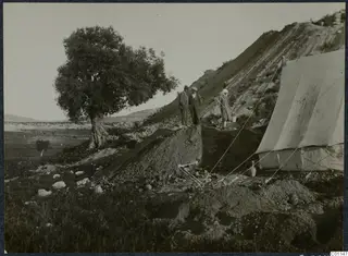 Two men and a woman standing on an excavated mound beside a tent and a large tree at Marion