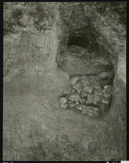 Excavated tomb entrance at Marion with a rubble of stones at its foot and grass on the bank above