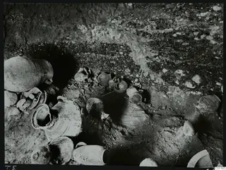 Marion T. 5 interior: pottery vessels and sherds lying on the floor of a tomb among stones and gravel.