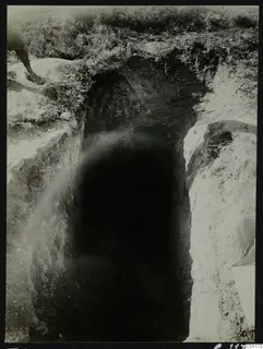 Tomb 11 at Marion during excavation, showing a dark opening cut into the ground with grass around the edges