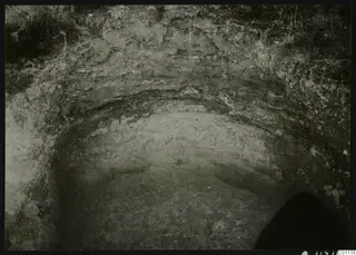 Excavated tomb cut at Marion seen from above, with layered soil walls and grass and roots along the top edge.