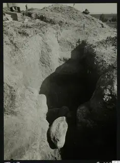 Excavation trench at Marion, Tomb 15, with a dark cut and large stones, people and grey boxes at the top edge.