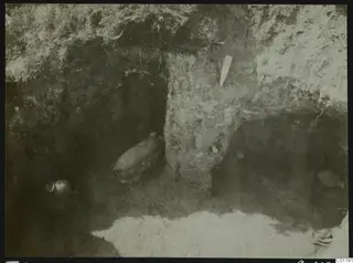Excavated tomb at Marion with pottery vessels lying in situ on the chamber floor and a pale tag in the earth wall.